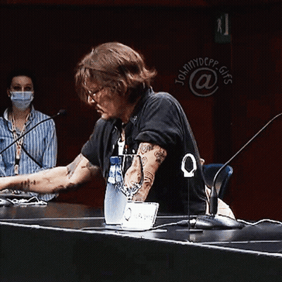 A Man Sits At A Table With A Laptop And A Bottle Of Water In Front Of A Sign That Says Johnny Depp GIF