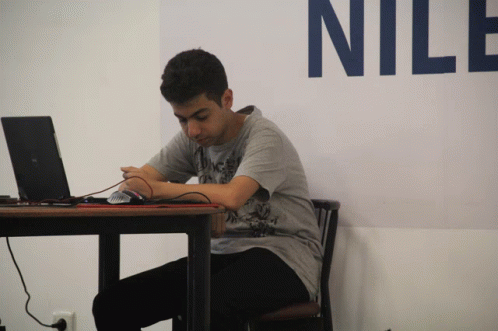 A Man Sits At A Table With A Laptop And A Mouse In Front Of A Wall That Says Nile GIF