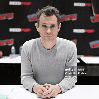 A Man Sits At A Table With His Hands Folded In Front Of A Wall That Says Reedpop GIF