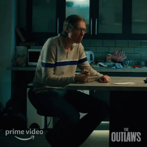 A Man Sits At A Table With The Words The Outlaws On The Bottom Right GIF