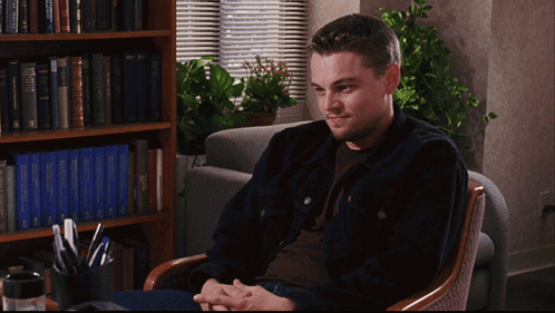 A Man Sits In A Chair In Front Of A Bookshelf With A Blue Book Titled " A Brief History Of The World " GIF