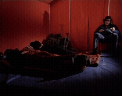A Man Sits In A Chair In Front Of A Red Wall While Another Man Lays On A Bed GIF