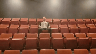 A Man Sits In A Row Of Red Seats In A Theater GIF