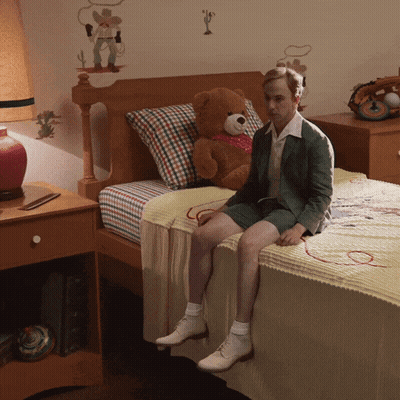 A Man Sits On A Bed With A Teddy Bear On It GIF