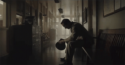 A Man Sits On A Bench In A Hallway Holding His Hat GIF