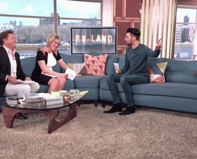 A Man Sits On A Blue Couch In Front Of A Sign That Says Ryland GIF
