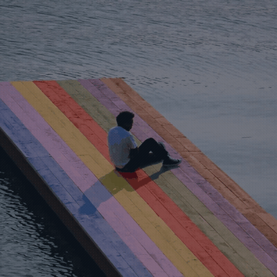 A Man Sits On A Colorful Wooden Dock Overlooking A Body Of Water GIF