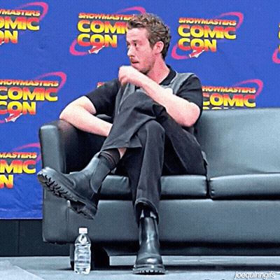 A Man Sits On A Couch In Front Of A Blue Wall That Says Showmasters Comic Con GIF