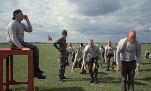 A Man Sits On A Red Bench Watching A Group Of Men Do Exercises In A Field GIF