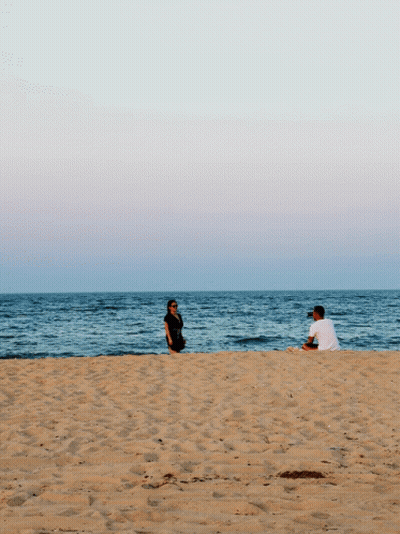 A Man Sits On The Beach While A Woman Stands On The Sand GIF