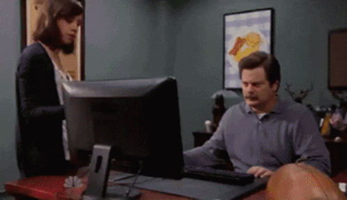 A Man Sitting At A Desk Using A Computer With A Woman Standing Behind Him GIF