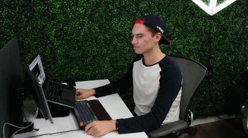 A Man Sitting At A Desk With A Laptop And A Hat That Says ' A ' On It GIF