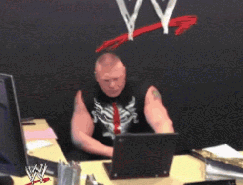 A Man Sitting At A Desk With A Laptop And A Wwe Logo On The Wall Behind Him GIF
