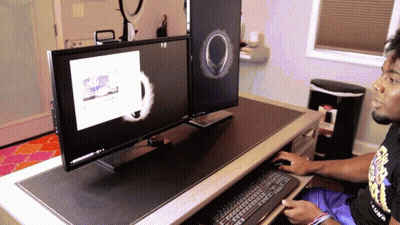 A Man Sitting At A Desk With Two Monitors And A Trash Can In The Background GIF
