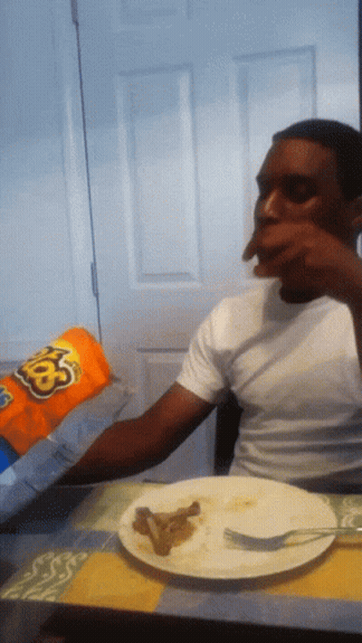 A Man Sitting At A Table Eating A Bag Of Cheetos GIF