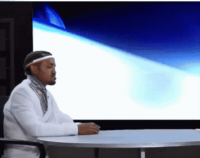 A Man Sitting At A Table In Front Of A Large Screen GIF