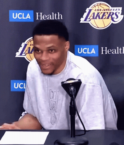 A Man Sitting At A Table In Front Of A Los Angeles Lakers Sign GIF