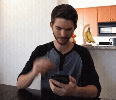 A Man Sitting At A Table Looking At A Cell Phone GIF