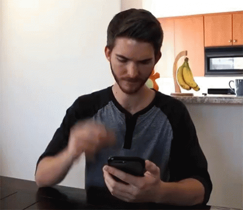 A Man Sitting At A Table Looking At A Cell Phone GIF