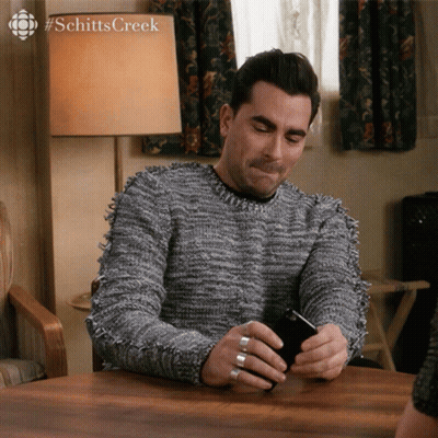 A Man Sitting At A Table Looking At A Cell Phone With #Schittscreek Written On The Bottom GIF