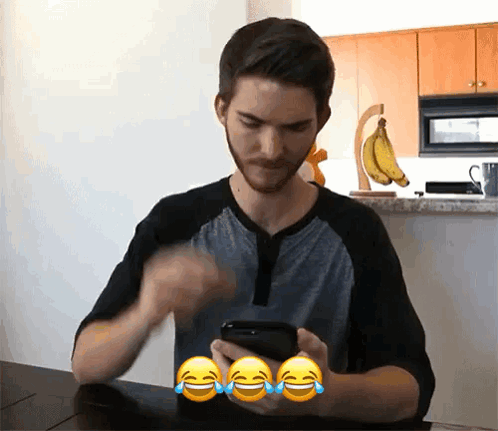 A Man Sitting At A Table Looking At His Phone With Three Laughing Emojis Around Him GIF