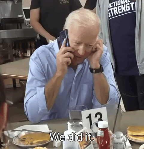 A Man Sitting At A Table Talking On A Cell Phone With A Sign That Says We Did It GIF