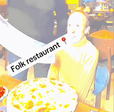 A Man Sitting At A Table With A Large Pizza And A Sign That Says Folk Restaurant GIF