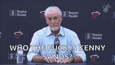 A Man Sitting At A Table With A Microphone And The Words Who The Fuck Is Kenny Sands Behind Him GIF