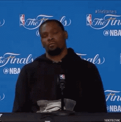 A Man Sitting At A Table With A Microphone In Front Of A Blue Background That Says The Finals On It GIF