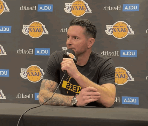 A Man Sitting At A Table With A Microphone In Front Of A Wall That Says Ajdu GIF