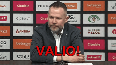 A Man Sitting At A Table With A Microphone In Front Of A Wall With Logos For Various Companies Including Valio GIF