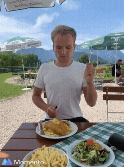A Man Sitting At A Table With A Plate Of Food And A Green Umbrella That Says Wieninger Bier GIF