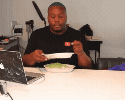 A Man Sitting At A Table With A Plate Of Food In Front Of A Laptop GIF