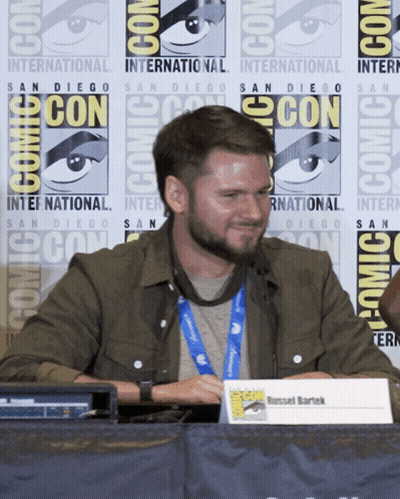 A Man Sitting At A Table With A Sign That Says Russell Bartek GIF