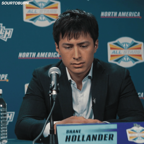 A Man Sitting At A Table With A Sign That Says Shane Hollander On It GIF