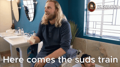 A Man Sitting In A Bathroom With The Words Here Comes The Suds Train On The Bottom GIF