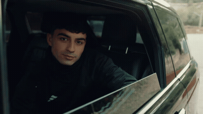 A Man Sitting In A Car With The Word Adidas On His Shirt GIF