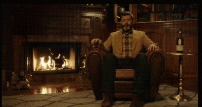A Man Sitting In A Chair In Front Of A Fireplace With A Bottle Of Wine On A Table GIF