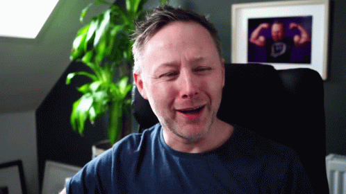 A Man Sitting In A Chair With A Picture Of A Man Flexing His Muscles On The Wall Behind Him GIF