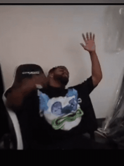 A Man Sitting In A Chair With His Hand In The Air Wearing A Shirt That Says ' Crysis ' On It GIF