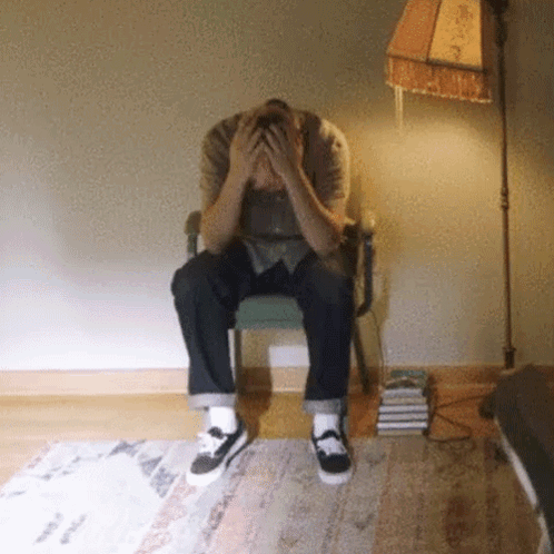 A Man Sitting In A Chair With His Hands On His Head GIF