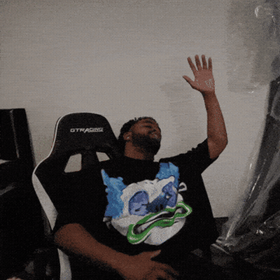 A Man Sitting In A Gtracing Chair With His Hand Up GIF