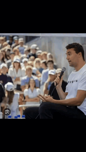 A Man Sitting In Front Of A Crowd Holding A Microphone And Wearing A T-Shirt That Says Reein GIF