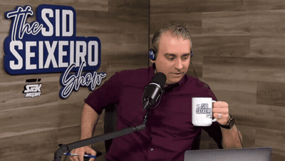 A Man Sitting In Front Of A Microphone With A Mug That Says The Sid Seixeiro Show GIF