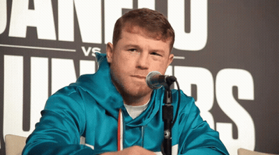 A Man Sitting In Front Of A Microphone With A Sign Behind Him That Says Juniors GIF