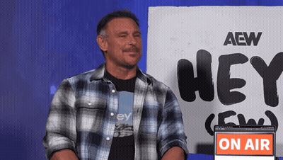 A Man Sitting In Front Of A Sign That Says Aew Hey GIF