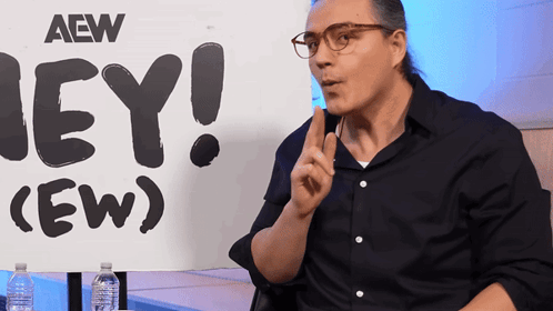 A Man Sitting In Front Of A Sign That Says Aew On It GIF