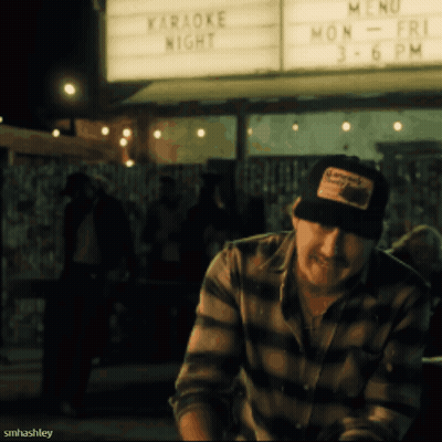 A Man Sitting In Front Of A Sign That Says " Karaoke Night " GIF