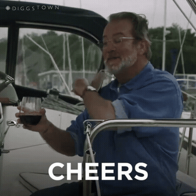 A Man Sitting On A Boat Holding A Glass Of Wine With The Word Cheers Behind Him GIF