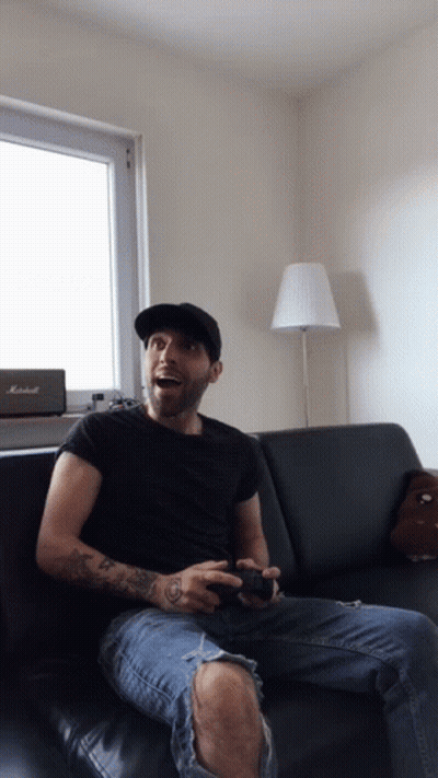 A Man Sitting On A Couch Playing A Video Game With A Marshall Speaker In The Background GIF